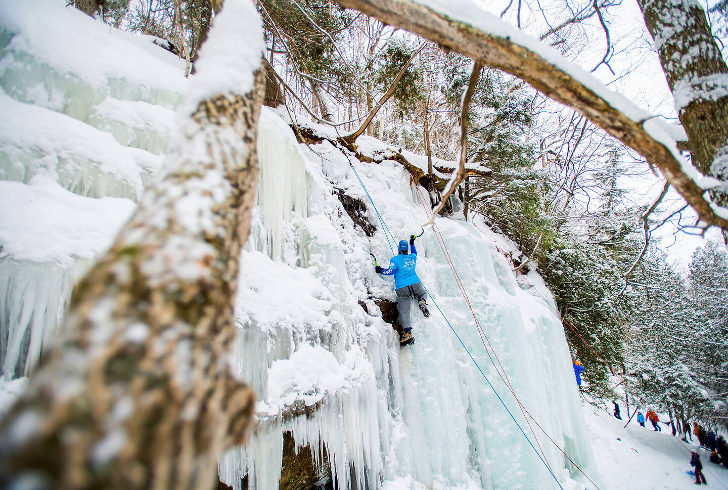 A person ascends a frozen waterfall using ice climbing gear, surrounded by snow-covered trees and a snowy trail with other climbers below.